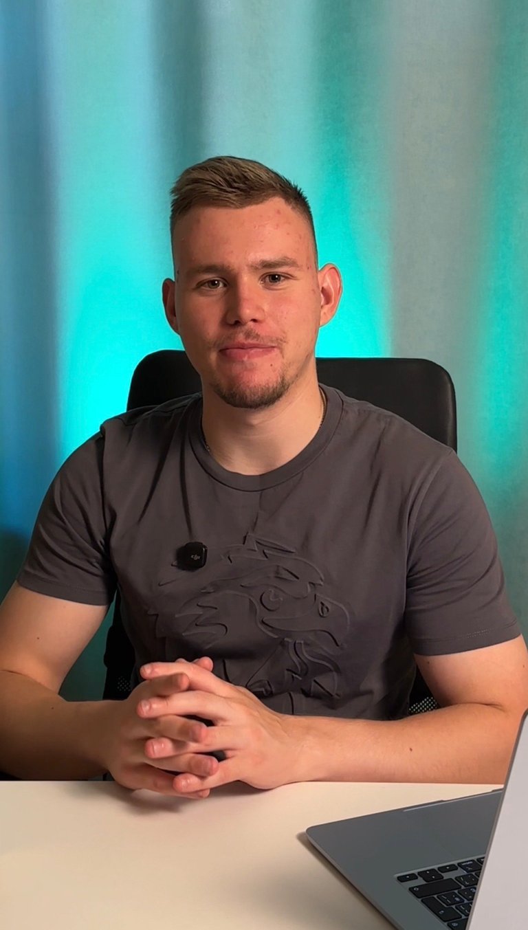 Man in dark t-shirt seated at desk with laptop, hands folded, against turquoise and blue gradient background